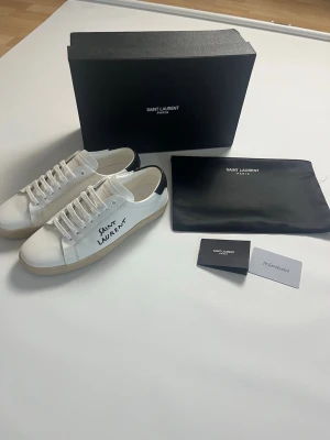 Saint Laurent vita sneakers i läder storlek 41 - Snygga vita sneakers från Saint Laurent i äkta läder med svart detalj på hälen och handskriven logga på sidan. Klassisk låg modell med vita snören och beige sula. Perfekta för dig som gillar stilrena och exklusiva skor. Skorna har storlek 41 och är helt nya och aldrig burna