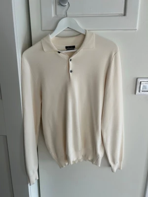 Krämfärgad stickad tröja Massimo Dutti - Stilren cremefärgad pikétröja från Massimo Dutti med lång ärm och tre knappar vid kragen. Tröjan har en mjuk, tunn stickning och ribbade muddar vid ärmslut och nederkant. Perfekt för dig som gillar en clean och tidlös look.