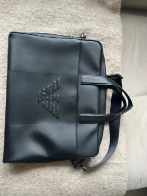 Svart datorväska från Emporio Armani - Stilren svart datorväska i slätt konstläder från Emporio Armani. Väskan har präglad logga på framsidan, dubbla handtag och justerbar axelrem. Perfekt för att bära laptop och viktiga prylar till skolan eller jobbet.