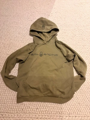 Olivgrön hoodie från Sail Racing - Olivgrön hoodie från Sail Racing med stor ficka framtill och logga tryckt över bröstet. Funkar för att slappa hemma och passar även väldigt bra med ljus blåa eller mörka jeans. Storlek 160 men jag tycker det passar bra för 165cm