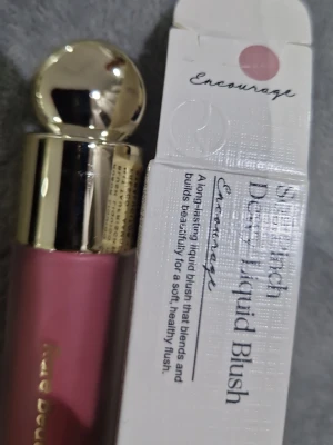 Rare Beauty Dewy Liquid Blush Encourage - Flytande rouge från Rare Beauty i nyansen Encourage, en mjuk rosa ton. Kommer i en elegant frostad glasflaska med guldig rund kork. Ger en naturlig, daggig finish och är lätt att blanda ut för ett fräscht resultat.