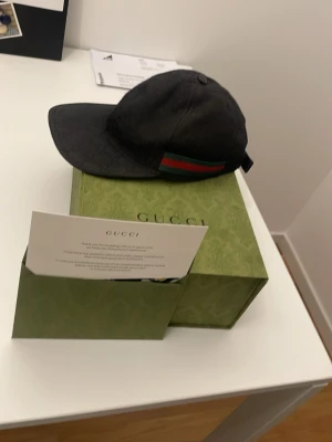 Svart keps från Gucci med logga - Snygg svart keps från Gucci med diskret GG-monogram över hela tyget och klassiska gröna och röda ränder på sidan. Kepsen har böjd skärm och justerbar rem bak. Perfekt accessoar för en trendig streetwear-look.