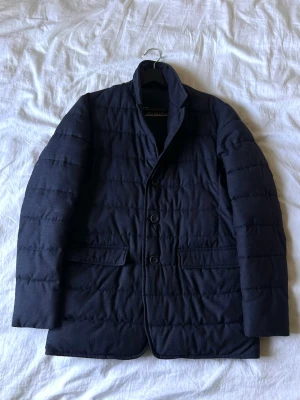Mörkblå pufferjacka från Herno - Snygg mörkblå pufferjacka från Herno med knäppning framtill och två fickor med lock. Jackan har en stilren design med lätt vaddering och hög krage. Perfekt för kalla dagar när du vill hålla stilen. Materialet är mjukt och ger en exklusiv känsla. Storleken är strl 48, passar S/M ungefär.