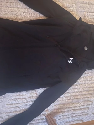 Svart vindjacka från Under Armour - Svart vindjacka från Under Armour med dragkedja framtill och huva. Jackan har diskret logga på bröstet och är tillverkad i lätt, vattenavvisande material med meshfoder för extra komfort. Perfekt för blåsiga dagar och sportiga outfits.