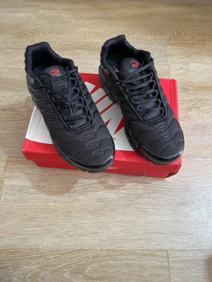 Nike Air Max Plus svarta sneakers - Svarta Nike Air Max Plus sneakers med röda detaljer och synlig Air Max-sula. Ovandelen är i mesh och syntet med vågformade linjer och klassisk Swoosh-logga på sidan. Skorna har en sportig vibe och är perfekta för dig som gillar streetwear.