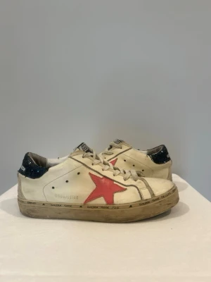 Golden Goose Dam Sneakers Med Rosa Stjärna - Snygga vita dam sneakers  från Golden Goose med en rosa stjärna på sidan och mörkblå glitterdetalj på hälen. Skorna har beige snören och slitna detaljer för en cool, vintage look. Tillverkade i läder och har textdetaljer runt sulan. Köpte i Rom Italien! Storlek US 9 - motsvarar 39.5-40. Skick 8/10.
