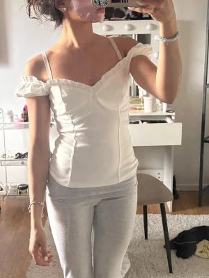 Vit omlott topp med volangdetaljer - Söt vit topp med omlottkänsla och volang längs urringningen. Axelbandslös look med smala axelband och lätt v-ringad hals. Figurnära passform och romantisk stil, perfekt för att lyfta vilken outfit som helst.
