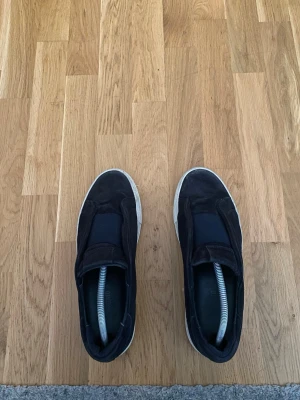Svarta Axel Arigatos i mocka - Stilrena svarta Axel Arigatos med ovandel i mocka och bred resår över vristen. Skorna har vit sula och minimalistisk design, perfekta för en clean och enkel look. Passar till det mesta och är lätta att ta på och av.