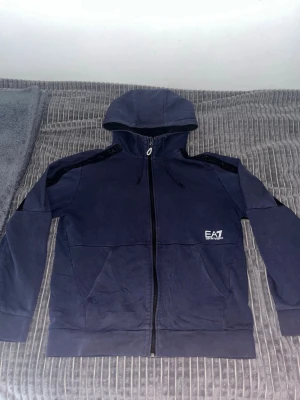 Mörkblå hoodie från EA7 Emporio Armani - Snygg mörkblå hoodie med dragkedja från EA7 Emporio Armani. Hoodien har huva, svarta detaljer på axlar och under armarna samt två fickor framtill. Tillverkad i mjukt bomullsmaterial och har logga på bröstet. Perfekt för en avslappnad och sportig stil.