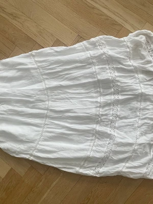 Vit långkjol med spetsdetaljer - Säljer en vit långkjol med fina spetsdetaljer och knyt i midjan. Kjolen har en luftig och bohemisk känsla, perfekt för varma dagar. Tillverkad i ett lätt och skönt material som ger en avslappnad look. Köpt i Spanien från hollister, knappt använd