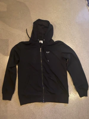 Svart hoodie från Comme des Garçons - Säljer en svart hoodie med dragkedja från Comme des Garçons Play. Den har huva med snörning den är strlk L men passar bäst för strlk M skriv om ni har frågor! 