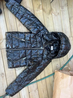 Svart glansig pufferjacka från Nelly - Snygg svart pufferjacka från Nelly i storlek 36. Jackan har glansigt material, huva och dragkedjor framtill samt två snedställda fickor med dragkedja. Perfekt för kalla dagar när du vill hålla stilen.