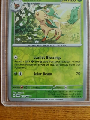 Pokémonkort Leafeon 001/167 Scarlet & Violet - Säljer ett Pokémonkort med Leafeon från Scarlet & Violet Paldean Fates, nummer 001/167. Kortet har grön bakgrund och motiv av Leafeon bland blommor. Kortet är gjort av papper och plast och ligger i ett genomskinligt skyddsfodral.