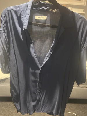 Blå skjorta från Jack & Jones - Säljer en stilren blå skjorta från Jack & Jones i storlek S. Skjortan har korta ärmar, klassisk krage och knappar framtill. Perfekt för en avslappnad men snygg look. Materialet känns lätt och luftigt, vilket gör den skön att bära under varmare dagar.
