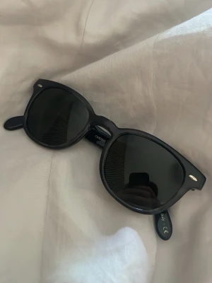 Svarta solglasögon från Oliver peoples  - Ny pris 3500. Klassiska svarta solglasögon från Oliver peoples med tidlös design och mörka glas. Bågarna är i plast och har en snygg, något fyrkantig form med små silvriga detaljer vid hörnen. Perfekta för soliga dagar och ger en cool vibe.