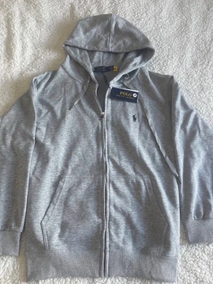 Grå hoodie från Polo Ralph Lauren - Snygg grå hoodie från Polo Ralph Lauren med dragkedja framtill och klassisk huva med dragsko. Broderad logga på bröstet och två fickor framtill. Tillverkad i mjukt bomullsmaterial och har ribbade muddar vid ärmslut och nederkant.