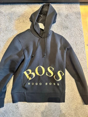 Svart hoodie från Hugo Boss - Svart hoodie från Hugo Boss med stor guld BOSS-logga framtill och justerbar dragsko i huvan. Tillverkad i mjukt bomullsmaterial och har ribbade muddar vid ärmslut och nederkant. Perfekt för en avslappnad och trendig stil.