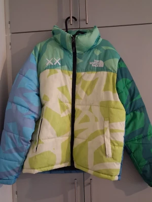 Flerfärgad vinter pufferjacka The North Face - Säljer en unik pufferjacka från The North Face med coolt blått, grönt och vitt mönster. Jackan har hög krage, dragkedja framtill och två fickor. Perfekt för dig som vill sticka ut i vinter och hålla dig varm med stil. Har använd jackan 2 gånger.