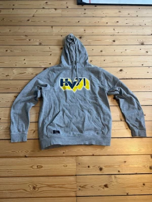 Grå HV71 hoodie med tryck - Säljer en grå hoodie från HV71 med stor gul och blå logga på bröstet. Hoodien har huva med dragsko och en klassisk magficka framtill. Tillverkad i mjukt bomullsmaterial, perfekt för dig som vill ha en sportig och avslappnad stil.