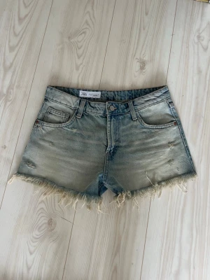 Lågmidjade Ljusblå jeansshorts från Zara - Säljer ett par lågmidjade ljusblå jeansshorts från Zara med fransig rå kant och slitna detaljer. Färgen är som en slags urtvättad blå. Användes bara ett fåtal gånger förra sommarn så fint skick!