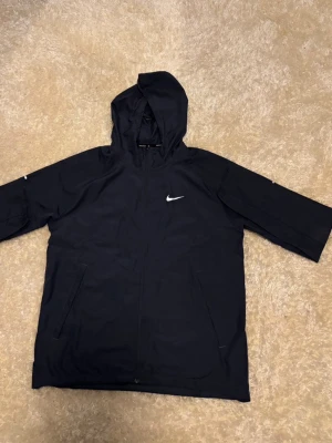 MÖRKBLÅ* Nike vindjacka med huva - Blå vindjacka från Nike med huva och dragkedja framtill. Jackan har två sidofickor med dragkedja och en vit Nike-logga på bröstet. Lätt och smidig modell, perfekt för blåsiga dagar. Materialet är syntetiskt och vattenavvisande.