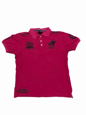 Rosa kortärmad polo Geographical Norway - Rosa kortärmad polo från Geographical Norway med pikékrage och knappar framtill. Tröjan har svarta broderade och tryckta detaljer på bröstet, ärmarna och ryggen, samt texten Royal Polo Club. Tillverkad i mjuk bomull för en bekväm känsla.