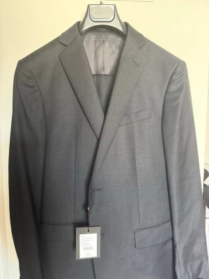 Grå kostymkavaj från John Varvatos - Snygg mörkgrå kostymkavaj från John Varvatos i klassisk modell. Kavajen har smala slag, två framfickor med lock och en bröstficka. Tillverkad i ull för en exklusiv känsla och stilren look. Perfekt för dig som vill ha en tidlös och elegant outfit. Fick den i present inför studenten men den passar inte mkg tyvärr. Den kostar 735€ ny pris
