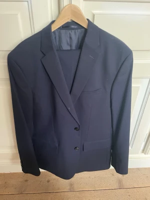 Mörkblå klassisk kostym - Snygg mörkblå kavaj med två knappar framtill och klassisk passform. Kavajen har slät yta, innerfickor och helfodrad insida. Perfekt till kostymbyxor eller jeans för en stilren look. Kostymbyxorna är i storlek 58 och jag har lagt in en bild på knäppningen. Kostymen är använd 1 gång och säljs eftersom att den har blivit för stor för mig. 