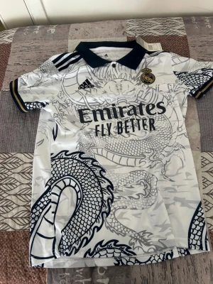 Real Madrid draktröja Adidas S - Säljer en vit Real Madrid matchtröja från Adidas med svart drakmönster och klubbmärke på bröstet. Tröjan har svart krage, korta ärmar och detaljer i guld och svart. Materialet är lätt och andas, perfekt för träning eller match.