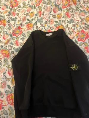 Svart sweatshirt från Stone Island - Svart sweatshirt från Stone Island med klassisk rund hals och ribbade muddar. Ikonisk logopatch på vänster ärm. Perfekt för en streetwear-look och enkel att matcha med jeans eller cargo. Tillverkad i mjukt bomullsmaterial.