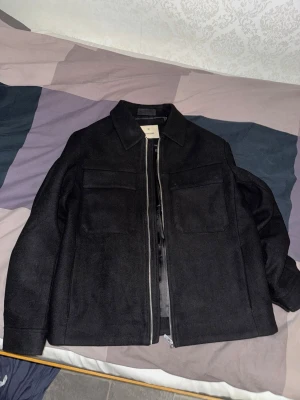 Overshirt från Jack & Jones - Säljer en svart overshirt från Jack & Jones med klassisk krage, två stora bröstfickor och dragkedja framtill. Jackan är fodrad med glansigt tyg på insidan och har en stilren, rak passform. Helt oanvänd. Säljs för att den är för stor. Fraktas endast och köparen står för frakt.