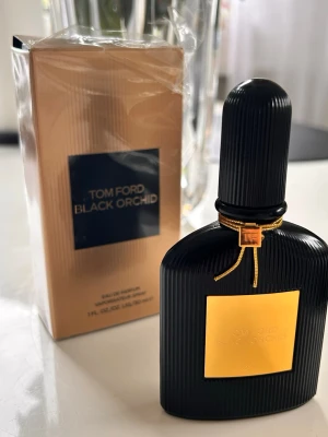 Tom Ford Black Orchid Eau de Parfum - Lyxig parfym från Tom Ford, Black Orchid, i en elegant svart flaska med guldiga detaljer och räfflad yta. Flaskan har en fyrkantig guldplatta framtill och ett dekorativt band runt halsen. Doften är ikonisk och passar dig som gillar exklusiva parfymer.