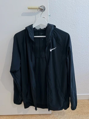 Svart Nike vindjacka med huva - Snygg svart vindjacka från Nike med huva och dragkedja. Jackan har Dri-FIT-material som andas och håller dig torr. Nike-logga i vitt på bröstet och praktiska fickor framtill. Perfekt för aktiva dagar och streetwear.