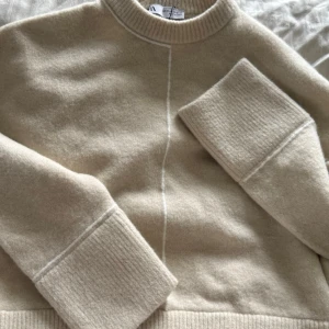 Beige stickad tröja från Zara - Säljer en beige stickad tröja från Zara med rund hals och breda ribbade muddar vid ärmslut och nederkant. Tröjan har en diskret söm framtill och är gjord i mjukt material, perfekt för lager-på-lager och chill dagar.