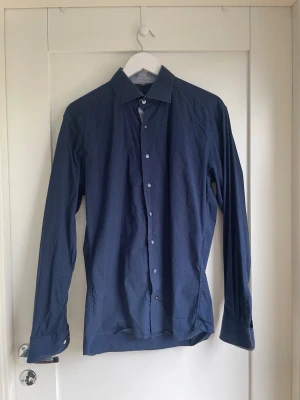 Mörkblå skjorta med småprickigt mönster - Snygg mörkblå skjorta med diskret småprickigt mönster och klassisk krage. Skjortan har långa ärmar och knäppning framtill. Perfekt för dig som gillar stilrena detaljer och en klassisk look.