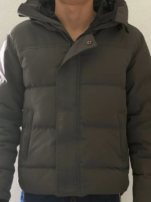 Grå  pufferjacka från Canada Goose MacMillen - Säljer en grå pufferjacka från Canada Goose med huva och diskret logotyp på ärmen. Jackan har två stora fickor framtill och stängs med dragkedja och knappslå. Tjockt vadderad för extra värme, perfekt för kalla vinterdagar.