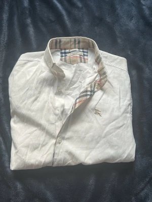 Vit skjorta från Burberry London - Klassisk vit skjorta från Burberry London med ikoniskt rutigt mönster på insidan av kragen och knappslån. Skjortan har knappar framtill och en liten broderad Burberry-logga på bröstet. Perfekt för en stilren och tidlös look.