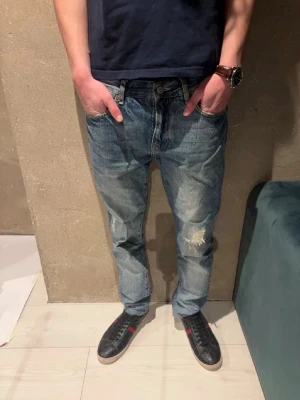 Jeans med slitningar - Säljer ett par blå jeans från Morris med rak passform och slitningar på ena benet. Jeansen har klassisk femficksdesign och en ljus tvätt med tydliga slitningar för en avslappnad look. Perfekta för dig som gillar en casual och trendig stil.