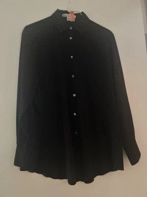 Svart skjorta från Acne Studios - Klassisk svart skjorta från Acne med rak passform och knappar framtill. Skjortan är tillverkad i ett mjukt, följsamt tyg som känns skönt mot huden. Perfekt att styla lager på lager eller bära som den är. Något oversized så passar en 36-38 i storlek. Nyss kemtvättad (se lapp)