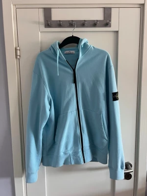 Ljusblå hoodie från Stone Island - Snygg ljusblå hoodie från Stone Island med dragkedja framtill och klassisk logopatch på vänster ärm. Hoodien har huva med dragsko och två stora fickor framtill. Tillverkad i mjuk bomull för skön känsla och avslappnad stil.