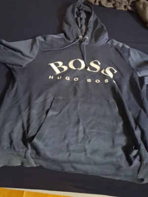 Mörk blå hoodie från Hugo Boss - Snygg mörk blå hoodie från Hugo Boss med stor vit BOSS-logga på bröstet och texten HUGO BOSS under. Klassisk huva med dragsko och stor magficka framtill. Tillverkad i mjukt bomullsmaterial som är skönt att bära. Perfekt för en avslappnad streetwear-look.