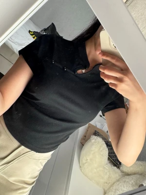 Svart ribbad t-shirt med cut out - Snygg svart ribbad t-shirt med korta ärmar och unik cut out-detalj vid halsen. T-shirten har en croppad passform och är perfekt för dig som gillar trendiga detaljer. Passar till både jeans och kjol.