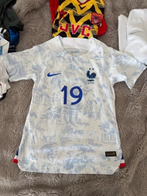Frankrike fotbollströja Nike - Säljer en oanvänd officiell Frankrike fotbollströja från Nike med nummer 19. Tröjan är vit med ljusblå mönster och har FFF-loggan med två stjärnor på bröstet. Tillverkad i ett lätt och ventilerande material, perfekt för match eller träning. Tight fit