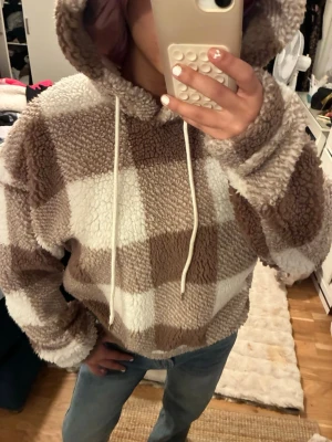 Rutig teddyhoodie i beige och vitt - Mysig teddyhoodie med stora rutor i beige och vitt. Hoodien har huva med vita dragsnören och är gjord i fluffigt, mjukt material som känns riktigt skönt mot huden. Storlek M men passar mig som bär XS. 