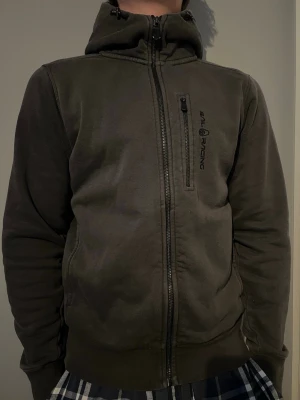 Mörkgrå hoodie från Sail Racing - Mörkgrå hoodie från Sail Racing med dragkedja framtill och en bröstficka med vertikal dragkedja. Märkets logga är broderad på bröstet. Hoodien har justerbar huva med snören och ribbade muddar vid ärmslut och nederkant. Perfekt för en avslappnad stil.