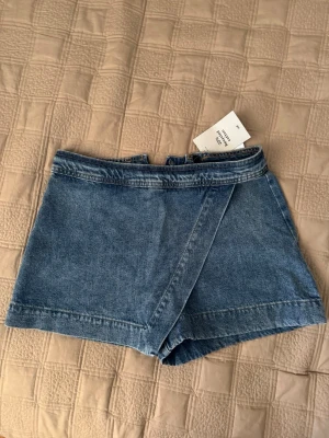 Blå denimshorts från H&M - Trendigt blå denimshorts från H&M med omlott-design framtill. Shortsen har klassisk jeanslook med synliga sömmar.