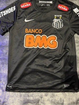 Svart Santos FC matchtröja Nike - Säljer en svart Santos FC matchtröja från Nike med Dri-FIT material. Tröjan har klubbmärke, sponsorloggor i vitt, rött och orange samt rund hals. Perfekt för fotbollsträning eller som supporterplagg.
