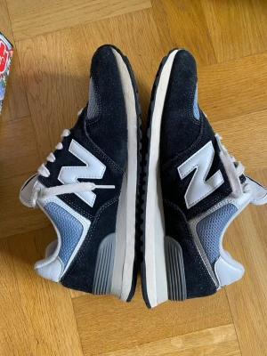 New Balance 574 svart/vit/blå storlek 41,5 - Snygga New Balance 574 sneakers i svart, vitt och blått. Ovandel i mocka och mesh med den klassiska N-loggan på sidan. Skön sula och retroinspirerad design som passar perfekt till streetwear och casual outfits.