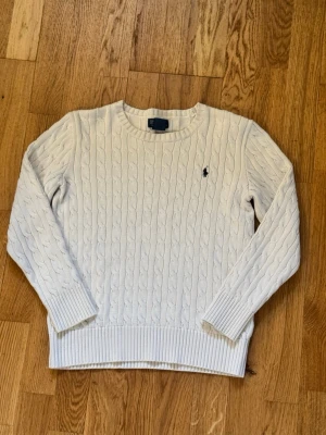 Vit kabelstickad tröja från Polo Ralph Lauren - Kabelstickad vit tröja från Polo Ralph Lauren med klassisk rund halsringning och den ikoniska mörkblå logotypen broderad på bröstet. Tröjan har ribbade muddar och nederkant, perfekt för en stilren look.