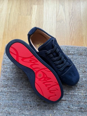 Mörkblå sneakers från Christian Louboutin - Snygga mörkblå sneakers i mocka från Christian Louboutin med klassisk röd sula och svarta skosnören. Skorna har en låg profil och stilren design som passar perfekt till streetwear eller en mer uppklädd look. Ikonisk logga på sulan och mjukt innerfoder.